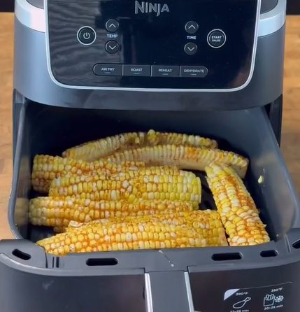 Six ears of corn cut in half are placed inside the basket of a Ninja air fryer. The corn appears to be seasoned and ready for air frying, with the air fryer sitting on a wooden surface.