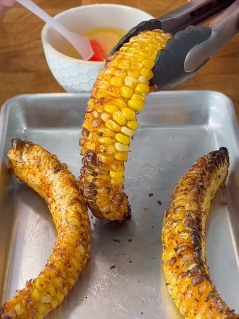 Three grilled corn cobs are on a metal tray. One is being held with tongs, while a hand uses a brush to apply melted butter from a small bowl in the background. The corn is charred and seasoned.