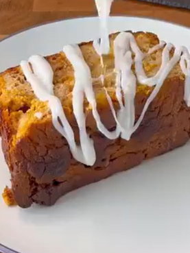 A slice of brown loaf cake on a white plate, with white icing being drizzled over the top.