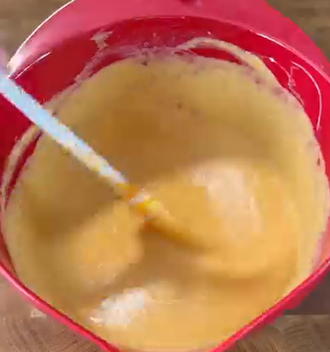 A close-up of a red mixing bowl filled with a thick, creamy yellow batter being stirred with a white spatula.
