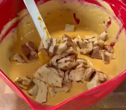 A red mixing bowl filled with thick yellow batter and chunks of chopped apples, being stirred with a white spatula.