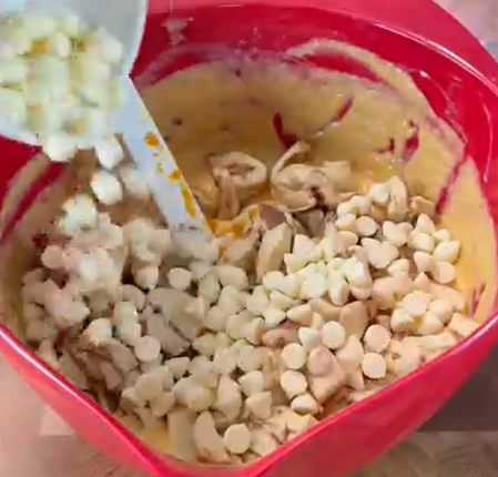 A red mixing bowl filled with batter, chopped apples, and white chocolate chips being added with a spatula.