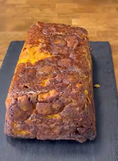 A rectangular loaf of marbled cake with a golden brown crust is placed on a black slate serving board, set on a wooden surface.