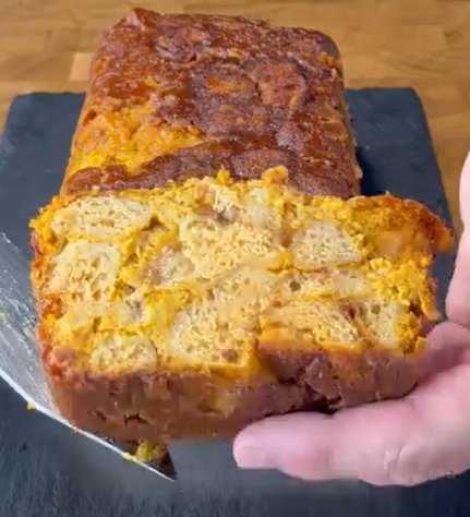 A hand holds a slice of baked bread pudding, showing its dense, moist interior with visible chunks of bread. The loaf rests on a dark surface with a golden-brown top.