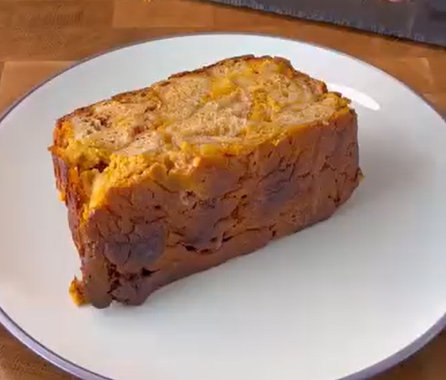 A thick slice of golden-brown bread pudding sits on a white plate with a silver rim, placed on a wooden surface.