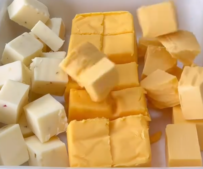 Cubes of white cheese and yellow cheese are arranged closely together in a container. The white cheese has small red and green specks, while the yellow cheese appears smoother and denser.