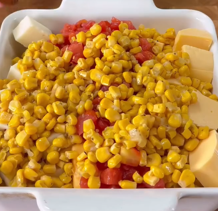 A white baking dish filled with corn kernels, diced tomatoes, and chunks of yellow and white cheese, arranged in separate sections.