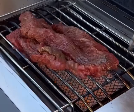 A large piece of raw beef is placed on a metal grill over a barbecue, with hot coals glowing underneath. The grill setup is clean and metallic, ready for cooking.