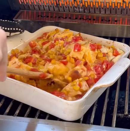 A hand stirs a cheesy casserole with tomatoes, corn, and tortilla pieces in a white baking dish placed on a grill. The dish looks hot and melty, with steam rising.