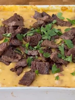 Chunks of grilled beef and fresh cilantro are scattered on top of creamy yellow cheese dip in a rectangular white dish.