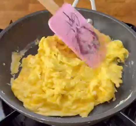 A pink spatula is stirring scrambled eggs in a non-stick frying pan on a stovetop.