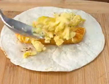 A tortilla on a wooden surface is topped with a piece of cooked sausage, while scrambled eggs are being added with a spoon.