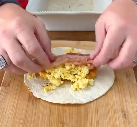 A person places a strip of cooked bacon on top of scrambled eggs and cheese on a tortilla, preparing to wrap it on a wooden cutting board.