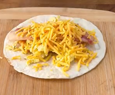 A flour tortilla on a wooden cutting board topped with scrambled eggs, cooked bacon, and shredded cheddar cheese.