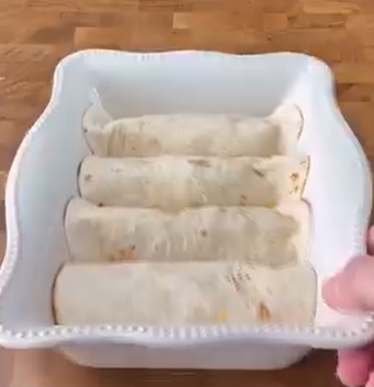 A hand holds a white, rectangular baking dish containing four rolled tortillas arranged neatly in a row on a wooden surface.