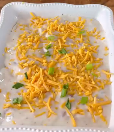 A close-up of a creamy dip topped with shredded cheddar cheese and chopped green onions, served in a white square dish with a decorative edge.