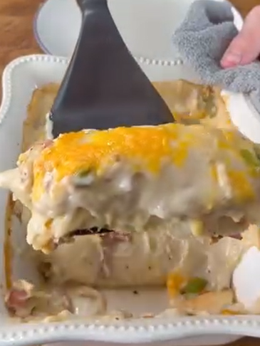 A close-up of a cheesy, creamy casserole being lifted from a white baking dish with a spatula. The casserole has a golden, melted cheese topping and visible layers inside.