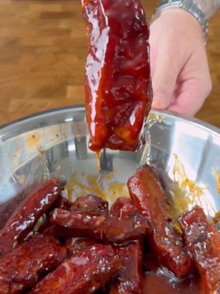 A hand holds up a glazed, saucy rib above a metal bowl filled with several similarly coated ribs, all covered in a shiny, sticky barbecue sauce.