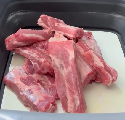 Several raw pork ribs are stacked together inside a rectangular container with a light-colored base. The ribs are uncooked, showing red meat and white fat.