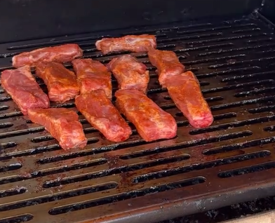 Several pieces of seasoned meat are cooking on a grill, spaced apart on black grates. The meat appears to be pork or beef, with visible reddish seasoning.