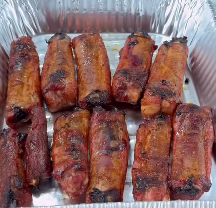 Twelve cooked pork ribs, with a slightly charred and glazed appearance, are arranged in two rows inside a rectangular aluminum tray.