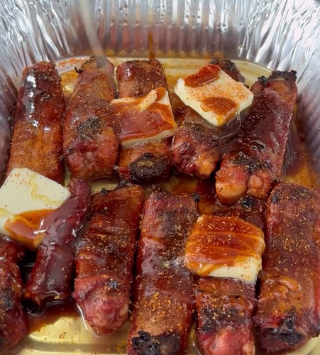 Several grilled, bacon-wrapped sausages are arranged in a foil tray, topped with pats of butter and drizzled with barbecue sauce. The sausages are seasoned and appear juicy and caramelized.