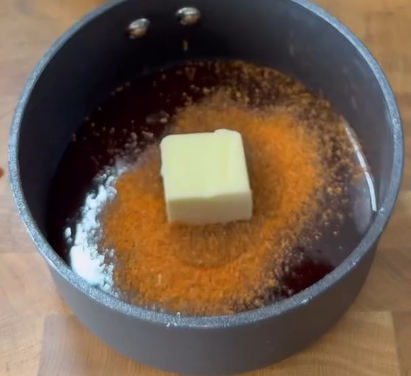 A black saucepan on a wooden surface contains dark liquid, a pat of butter in the center, and a sprinkling of reddish-brown seasoning around the butter.