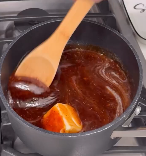 A wooden spoon stirs a dark brown sauce with a melting pat of butter in a black saucepan on a stovetop.