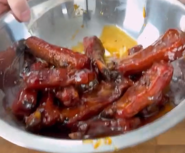 A large metal bowl filled with glazed, saucy pork ribs being held up, with a hand visible at the edge of the bowl. The ribs are coated in a rich reddish-brown sauce.