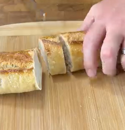 A hand reaching for a sliced baguette on a wooden cutting board.