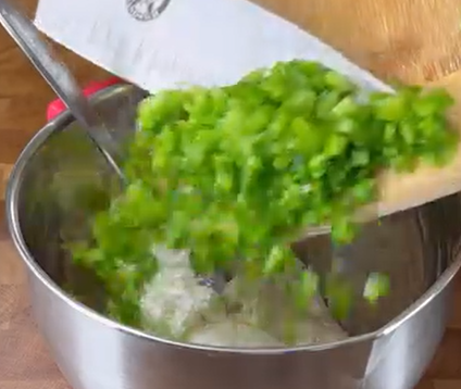 A person is adding chopped green bell peppers from a cutting board into a stainless steel mixing bowl that contains some white ingredients. A spoon is visible inside the bowl.
