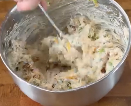 A hand stirs thick, chunky batter with a spoon in a large metal mixing bowl on a wooden surface.