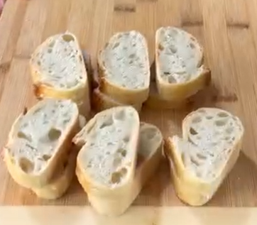 Six slices of crusty baguette bread are arranged on a wooden cutting board, displaying their airy, open crumb texture.