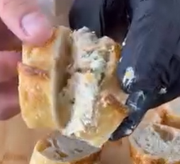 A close-up of hands breaking open a piece of bread stuffed with a creamy filling. One hand wears a black glove, while the other is bare.