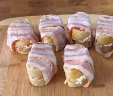 Six pieces of bread rolls filled with cream cheese and wrapped in strips of bacon are arranged on a wooden cutting board.