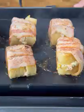 Four rectangular pieces of food wrapped in bacon strips are cooking on a black grill. The bacon is partly cooked and the filling inside appears to include cheese. Steam is rising from the hot surface.