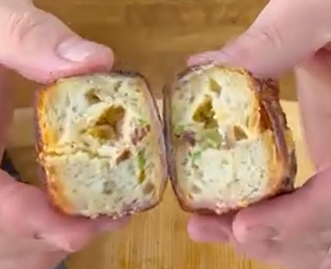 Two hands hold and pull apart a square piece of bread or pastry, revealing a soft, airy inside with small chunks of green and orange ingredients. The surface looks golden brown.
