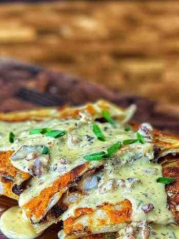 Close-up of crispy quesadillas topped with a creamy sauce containing diced mushrooms and garnished with chopped green onions, served on a wooden surface.