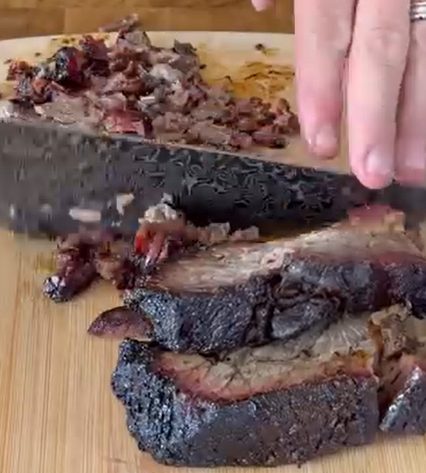 A hand slices smoked brisket on a wooden cutting board. Several thick slices with dark, charred edges are shown, and chopped pieces of meat are gathered near the knife.