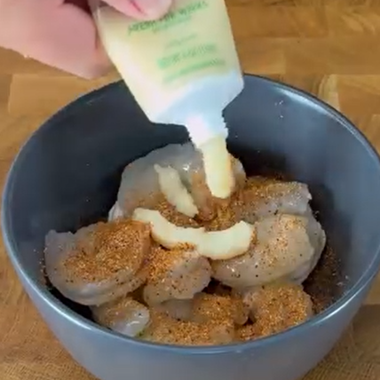 A hand squeezes a bottle of creamy sauce over raw shrimp coated with seasoning in a dark bowl on a wooden surface.