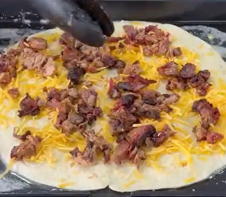 A gloved hand sprinkles chunks of cooked meat over shredded cheese on a round, uncooked pizza dough.