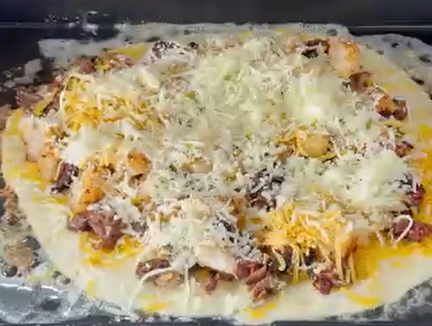 A close-up of an uncooked pizza topped with shredded cheese, pieces of chicken, and what appears to be shredded beef, all on a round pizza dough, ready to be baked.