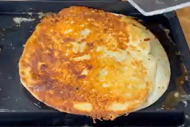 A large, round flatbread with a golden-brown, crispy surface is cooking on a black griddle. A spatula is positioned near the edge, ready to flip or remove the bread.