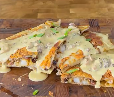 Sliced quesadillas filled with cheese and vegetables are topped with a creamy mushroom sauce and garnished with chopped green onions, served on a wooden board.