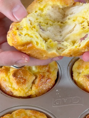 A person holds a cheesy, fluffy muffin broken in half, revealing a soft interior. More muffins are in a metal muffin tin below.