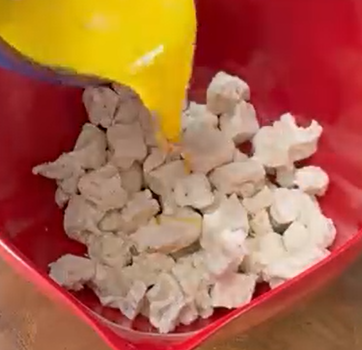 A yellow liquid is being poured from a container onto chunks of cooked chicken in a red mixing bowl.