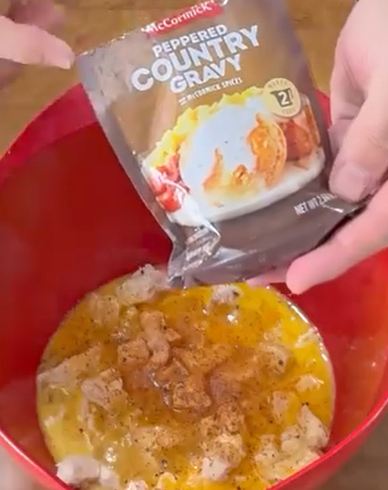 A person pours a packet of McCormick Peppered Country Gravy mix into a red bowl containing whisked eggs and cubed bread pieces.