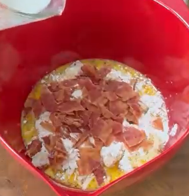 A red mixing bowl filled with flour, crumbled bacon, and beaten eggs, with milk being poured in from a glass measuring cup.