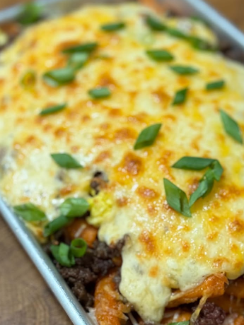 A close-up of a baked casserole topped with melted, golden-brown cheese and garnished with chopped green onions, on a metal baking tray.