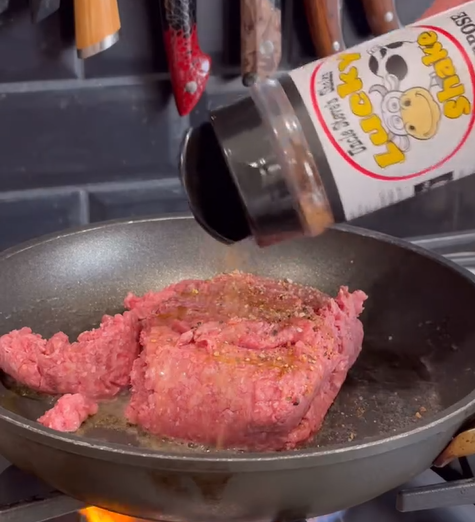 Ground beef cooks in a frying pan on a stove while a hand sprinkles seasoning from a container labeled Lucky Shake over the meat. Knives hang on a magnetic strip in the background.